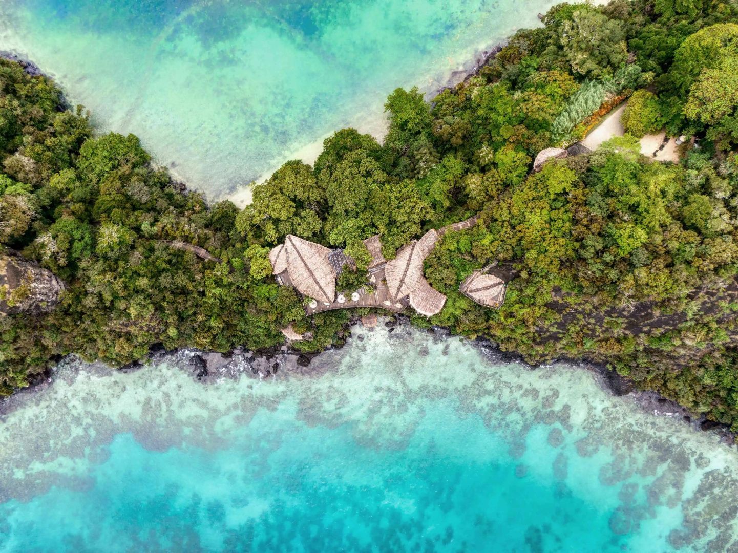 Luftaufnahme von Reetdachvillen auf einer grünen Landzunge umgeben von türkisblauem Meer.