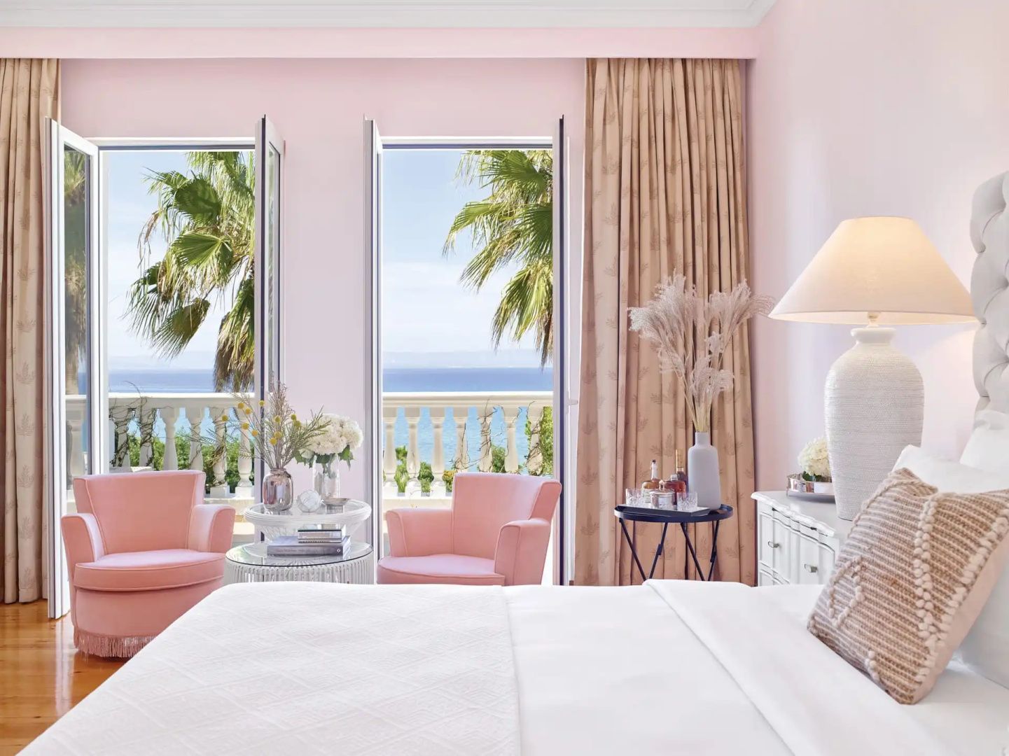 Rosa gestaltetes Hotelzimmer mit Doppelbett, rosa Sesseln und Balkontüren mit Blick auf das Meer.