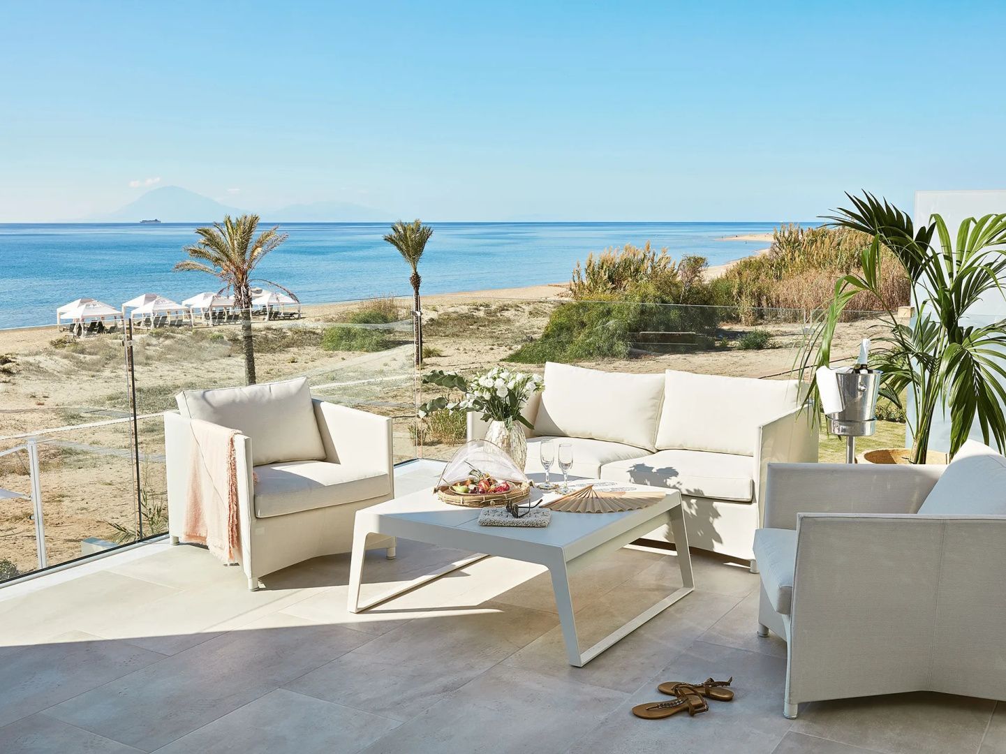 Weißes Loungeset auf einer Terrasse mit freiem Blick auf den Sandstrand und das blaue Meer.