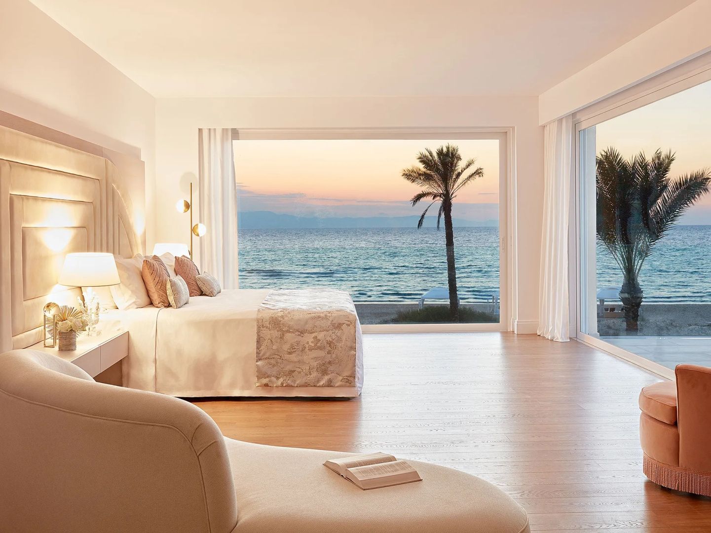 Hell eingerichtetes Schlafzimmer mit Doppelbett, Liege und Blick auf das Meer bei Sonnenuntergang.