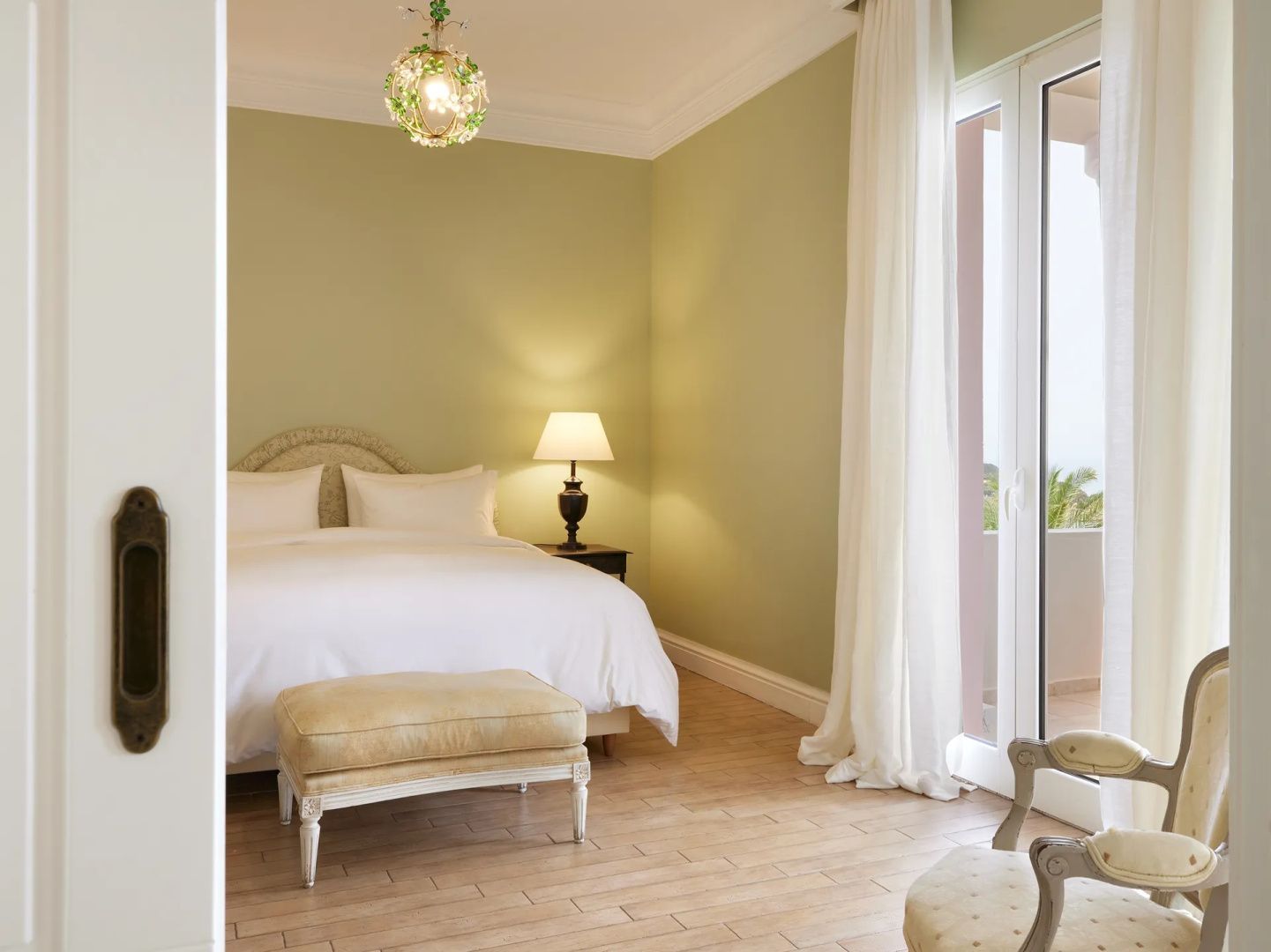 Schlafzimmer mit grün gestrichenen Wänden, weißem Doppelbett, Polsterbank und Balkonzugang.