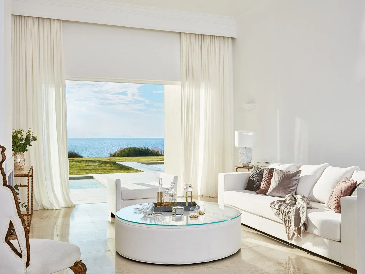 Heller Wohnbereich mit weißem Sofa, rundem Glas-Couchtisch und Durchgang zum Außenpool und Meer.