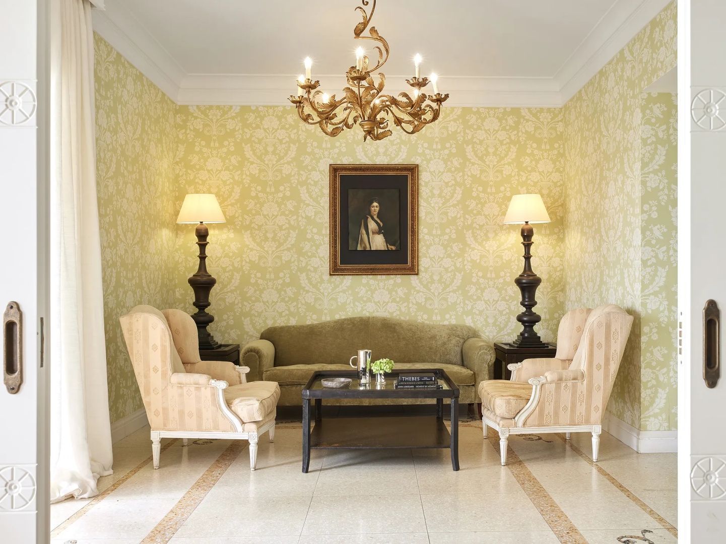 Wohnzimmer mit gemusterter Tapete, einem grünen Sofa, zwei Sesseln und Kronleuchter an der Decke.