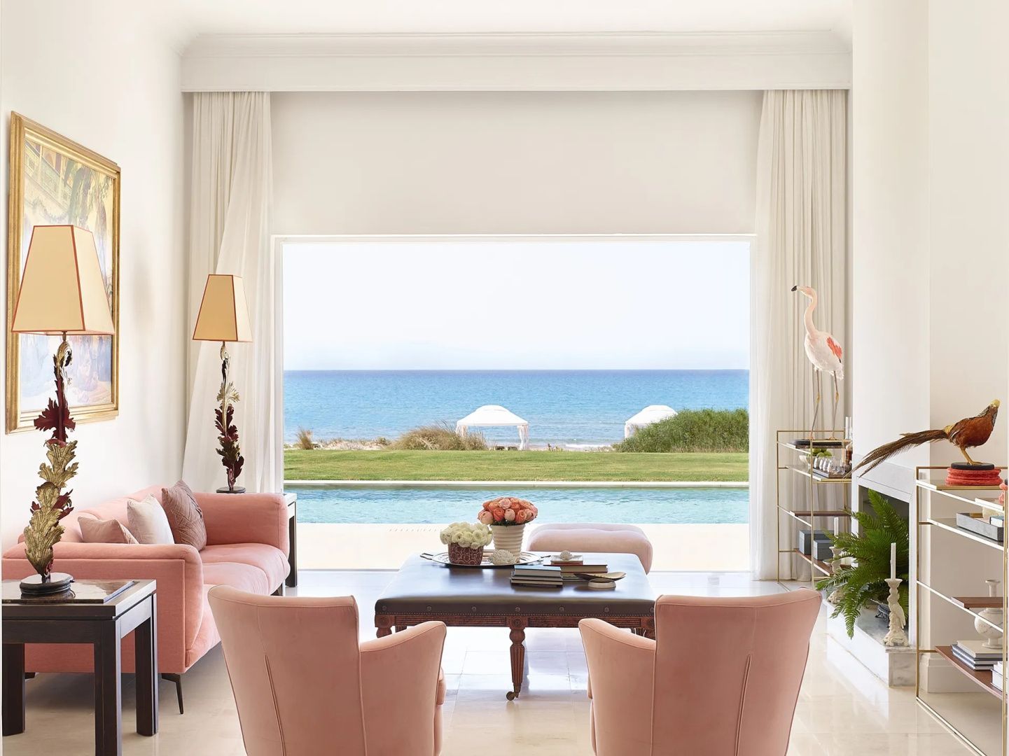 Wohnraum mit rosa Sesseln und großer Fensterfront mit Blick auf einen Pool, Rasen und den Strand.