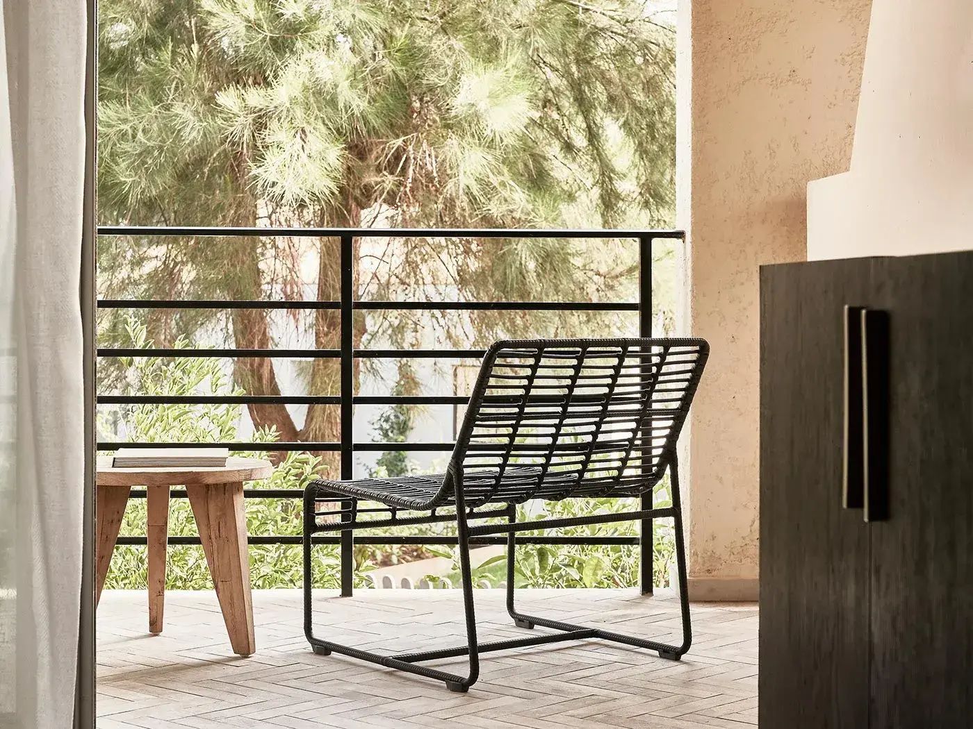 Balkon mit einem schwarzen Stuhl aus Geflecht und Holztisch vor grünen Bäumen.