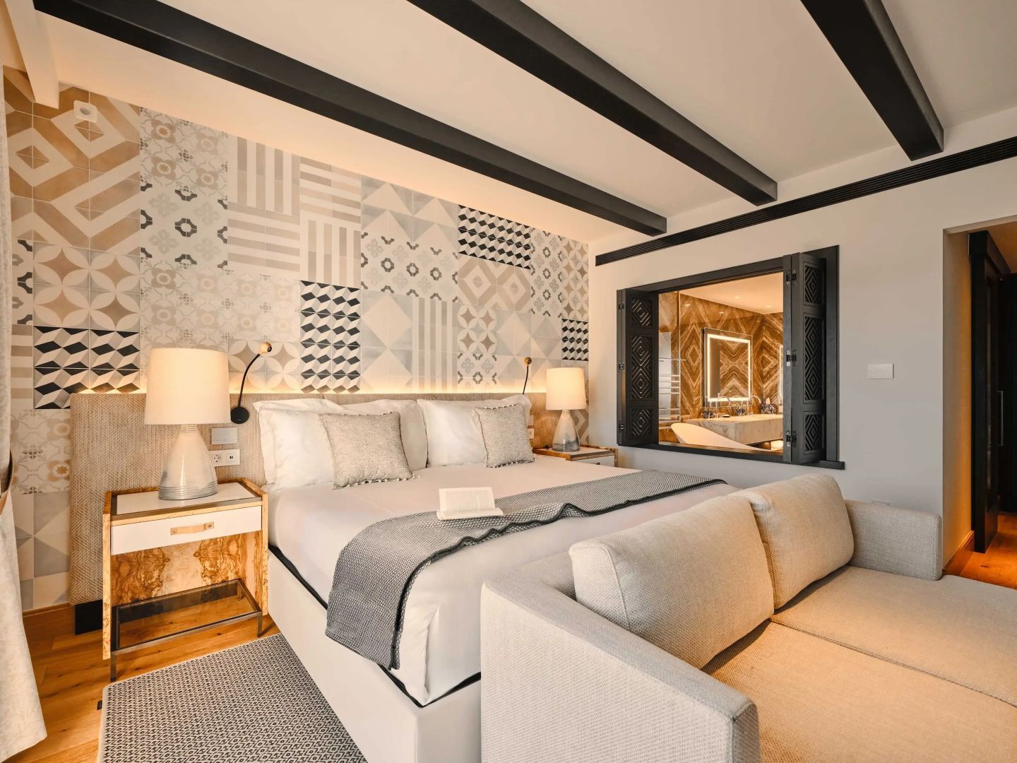 Doppelbett mit gemusterter Fliesenwand, Holzdeckenbalken und Blick ins Badezimmer.