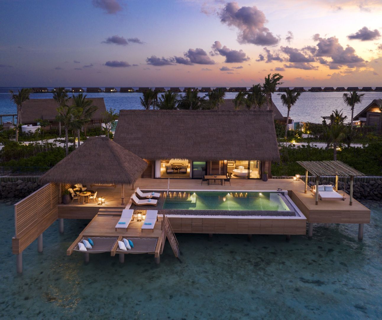 Wasservilla bei Dämmerung mit beleuchtetem Pool, Holzdeck und Reetdach über dem Meer.
