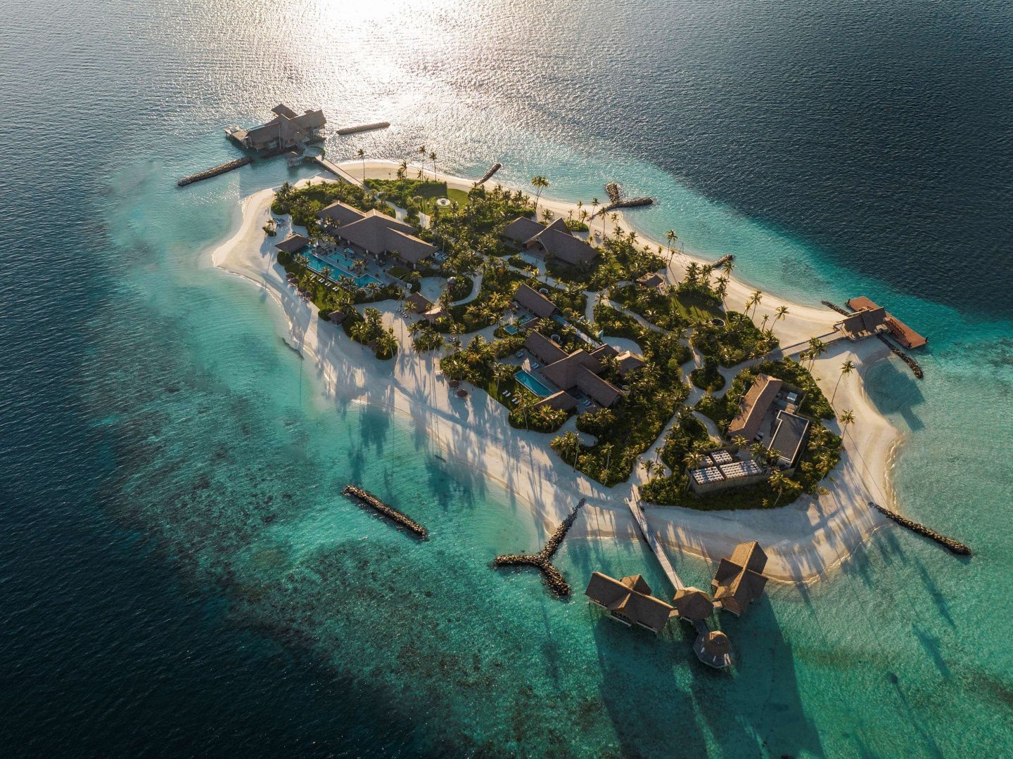 Luftaufnahme einer Insel mit üppiger Vegetation, Sandstrand und Villen im flachen Meerwasser.