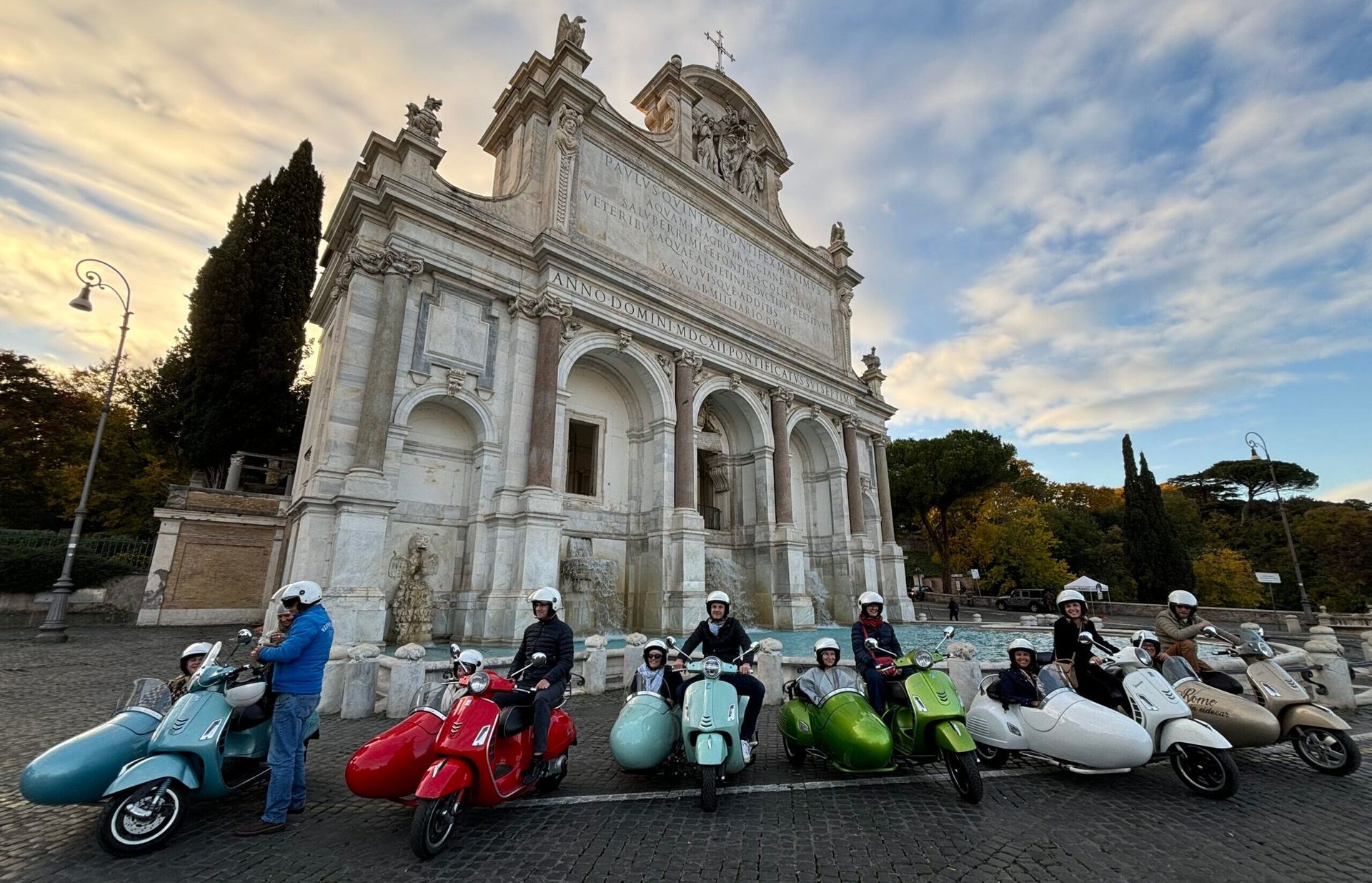 Gruppe von Leuten auf farbigen Vespas vor historischem Brunnen