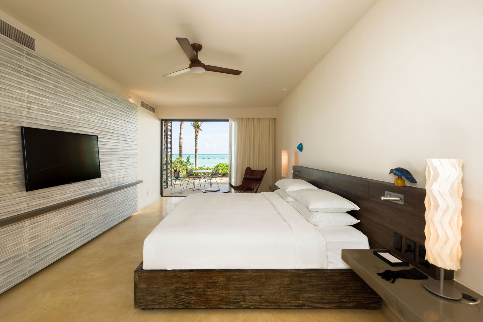 Helles Hotelzimmer mit großem Doppelbett, Deckenventilator und Blick auf die Terrasse und das Meer.