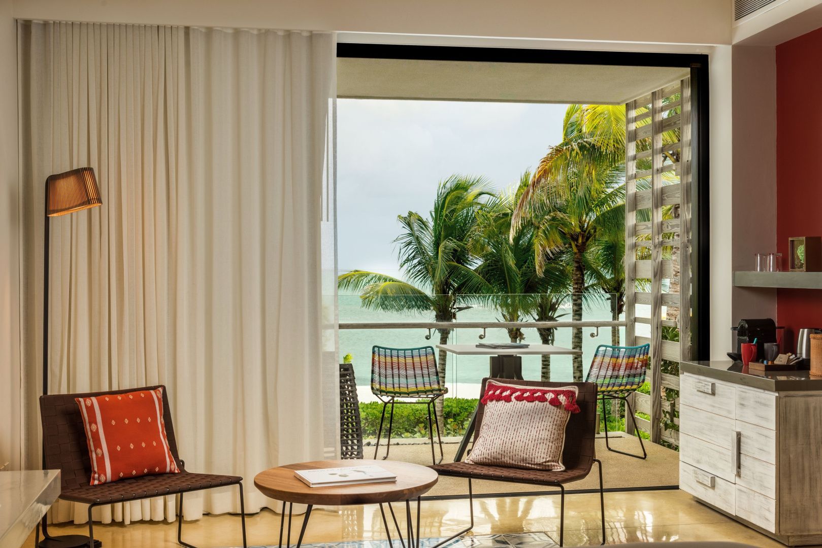 Blick aus dem Zimmer über die Terrasse mit bunten Stühlen auf Palmen und das Meer.