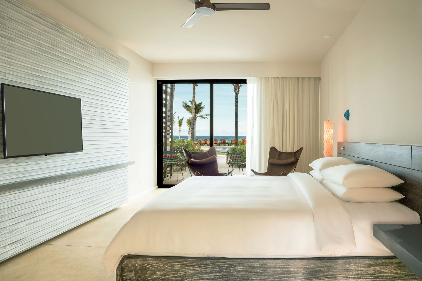 Helles Hotelzimmer mit großem Doppelbett und Blick durch die Glasfront auf die Terrasse und das Meer.
