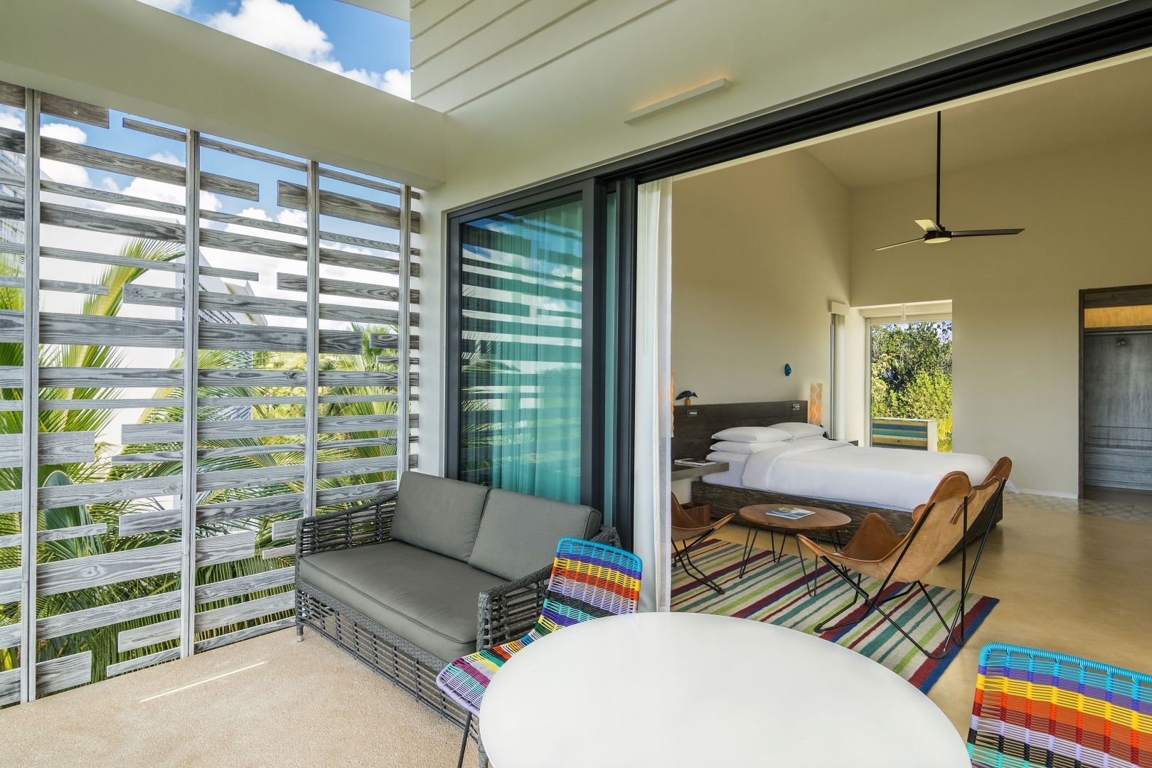 Überdachte Terrasse mit grauer Couch, buntem Stuhl und geöffneter Schiebetür zum Schlafzimmer.