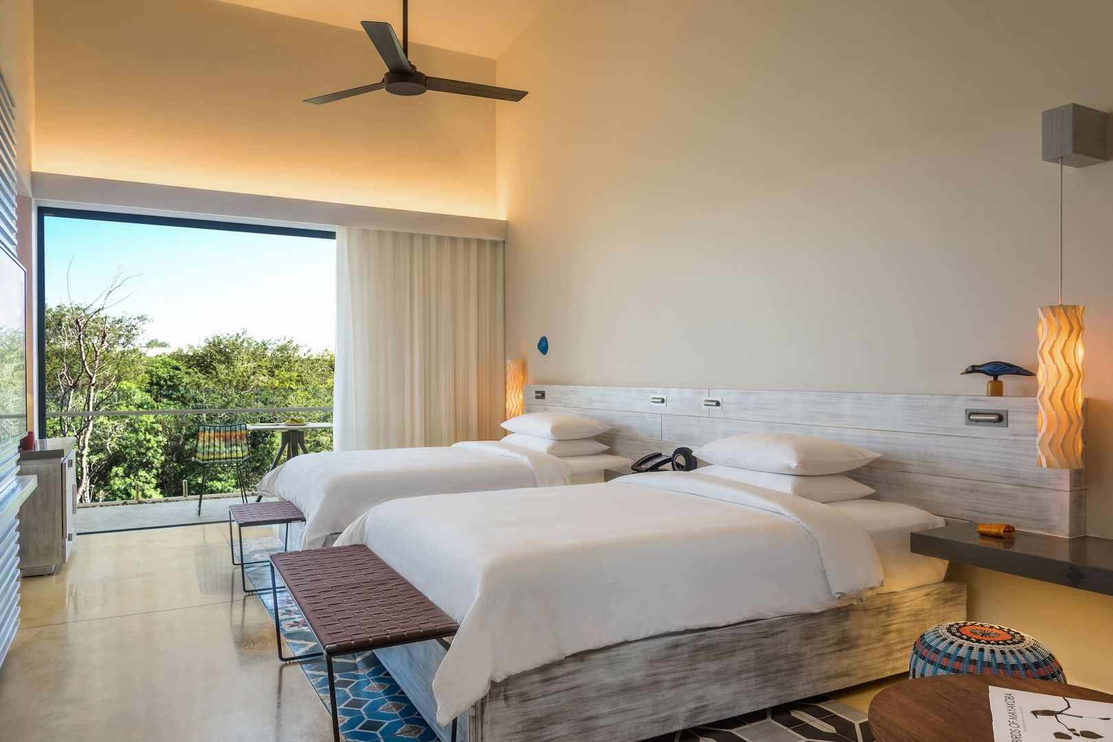 Helles Hotelzimmer mit zwei Doppelbetten, Deckenventilator und Zugang zum Balkon im Grünen.