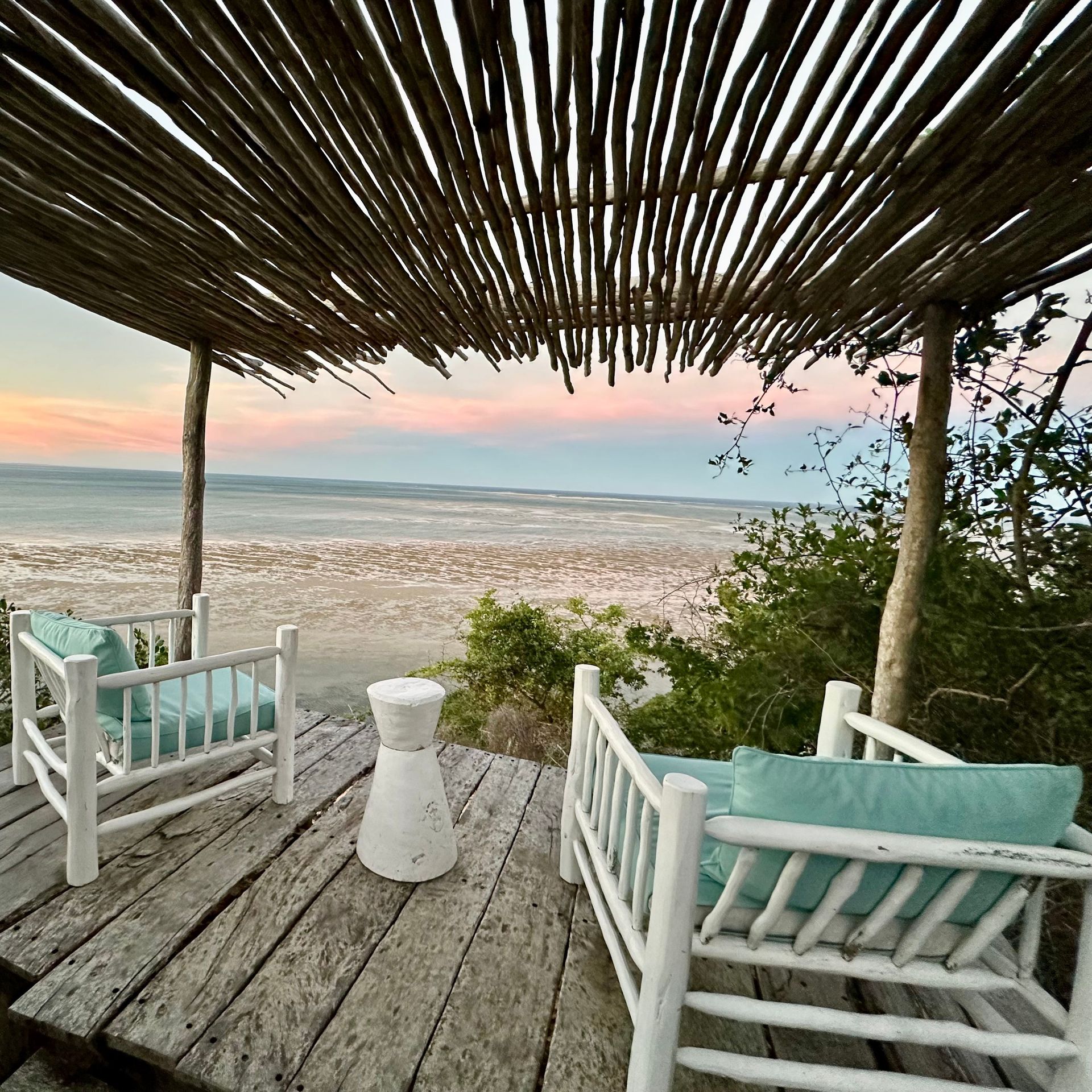 Zwei Holzstühle mit Kissen unter Pergola mit Blick auf Sandstrand bei Sonnenuntergang