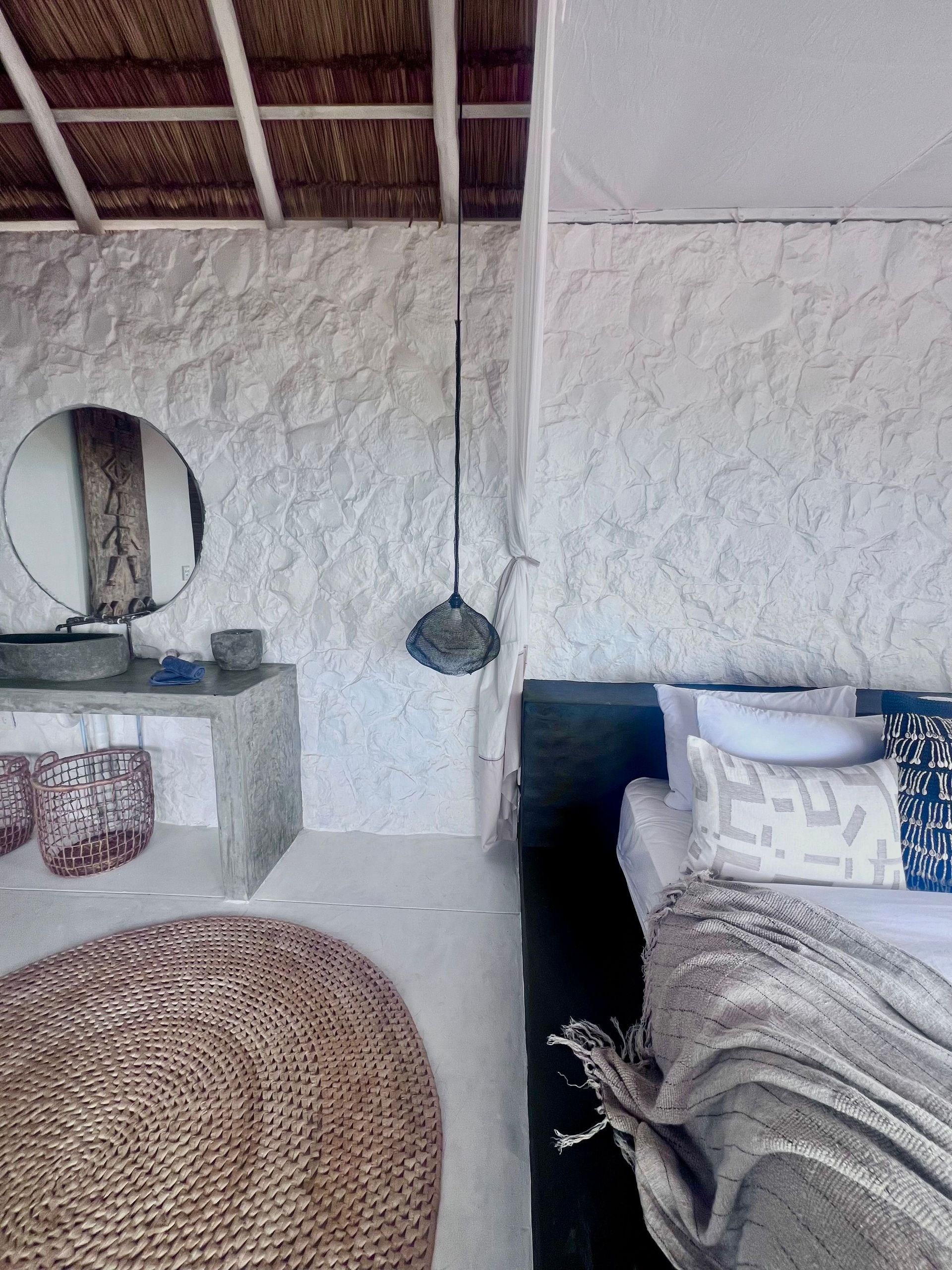 Schlafzimmer mit weißer Steinwand, Bett und runder, geflochtener Matte am Boden.