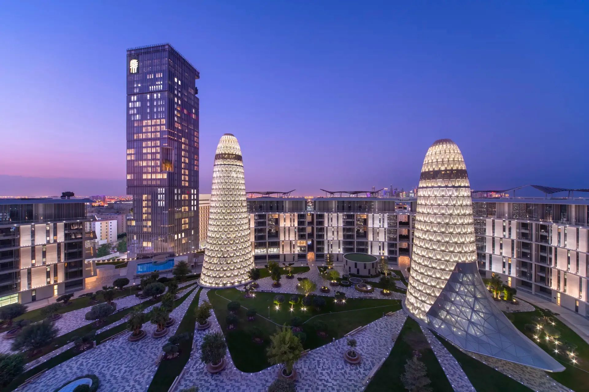  Abendaufnahme des Banyan Tree Doha Resorts im futuristischen Doha Oasis-Komplex, mit beleuchteten, kegelförmigen Glastürmen, modernem Hochhaus und elegantem Innenhof mit begrünten Wegen und luxuriöser Architektur unter violett-blauem Himmel.