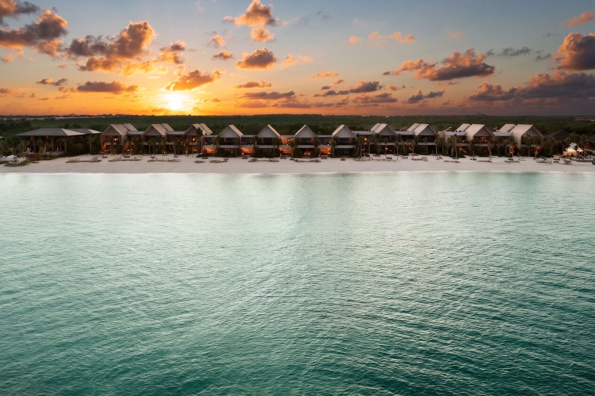 Luxuriöse Strandvillen des Banyan Tree Mayakoba Resorts in Mexiko bei Sonnenuntergang, mit ruhigem türkisfarbenem Meer im Vordergrund und dramatisch gefärbtem Himmel im Hintergrund.