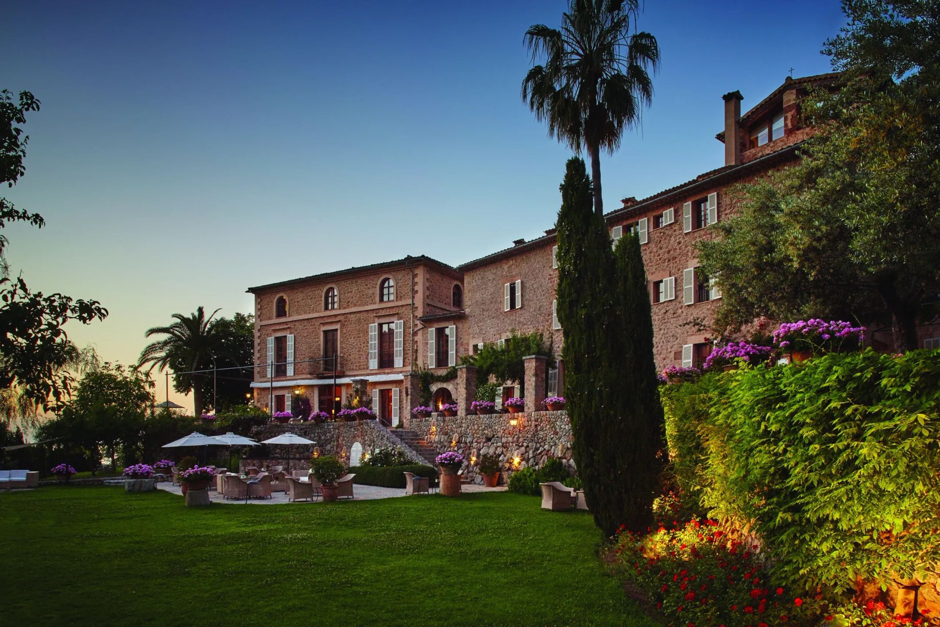Historische Villa in einer üppigen Gartenlandschaft bei Sonnenuntergang, umgeben von Palmen und Blumen.