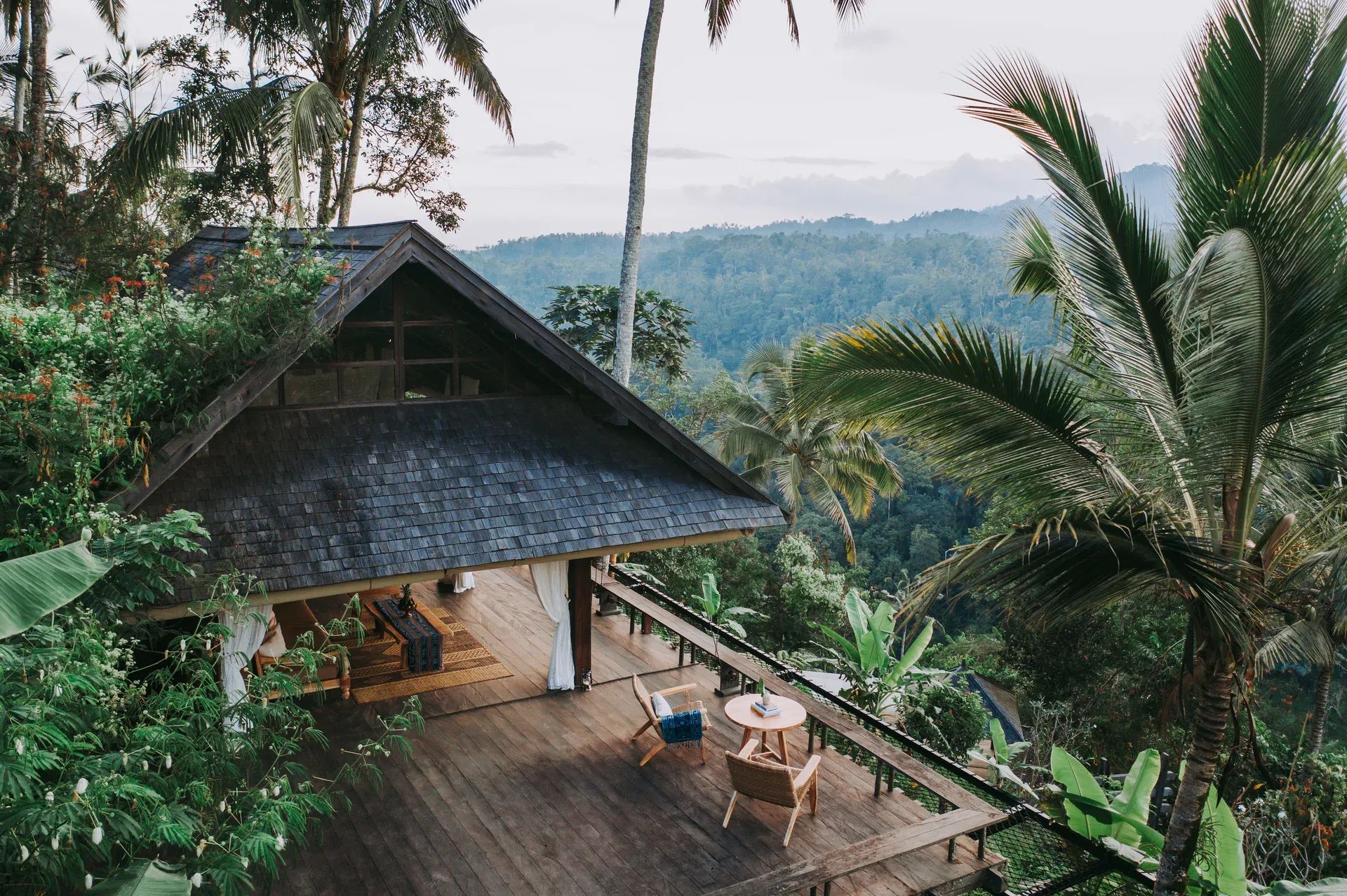 Offene Luxusvilla im Dschungel des Buahan, a Banyan Tree Escape Resorts auf Bali, mit Holzdeck, traditionellen Materialien und atemberaubendem Blick auf dichte tropische Wälder und Palmen unter einem leicht bewölkten Himmel.