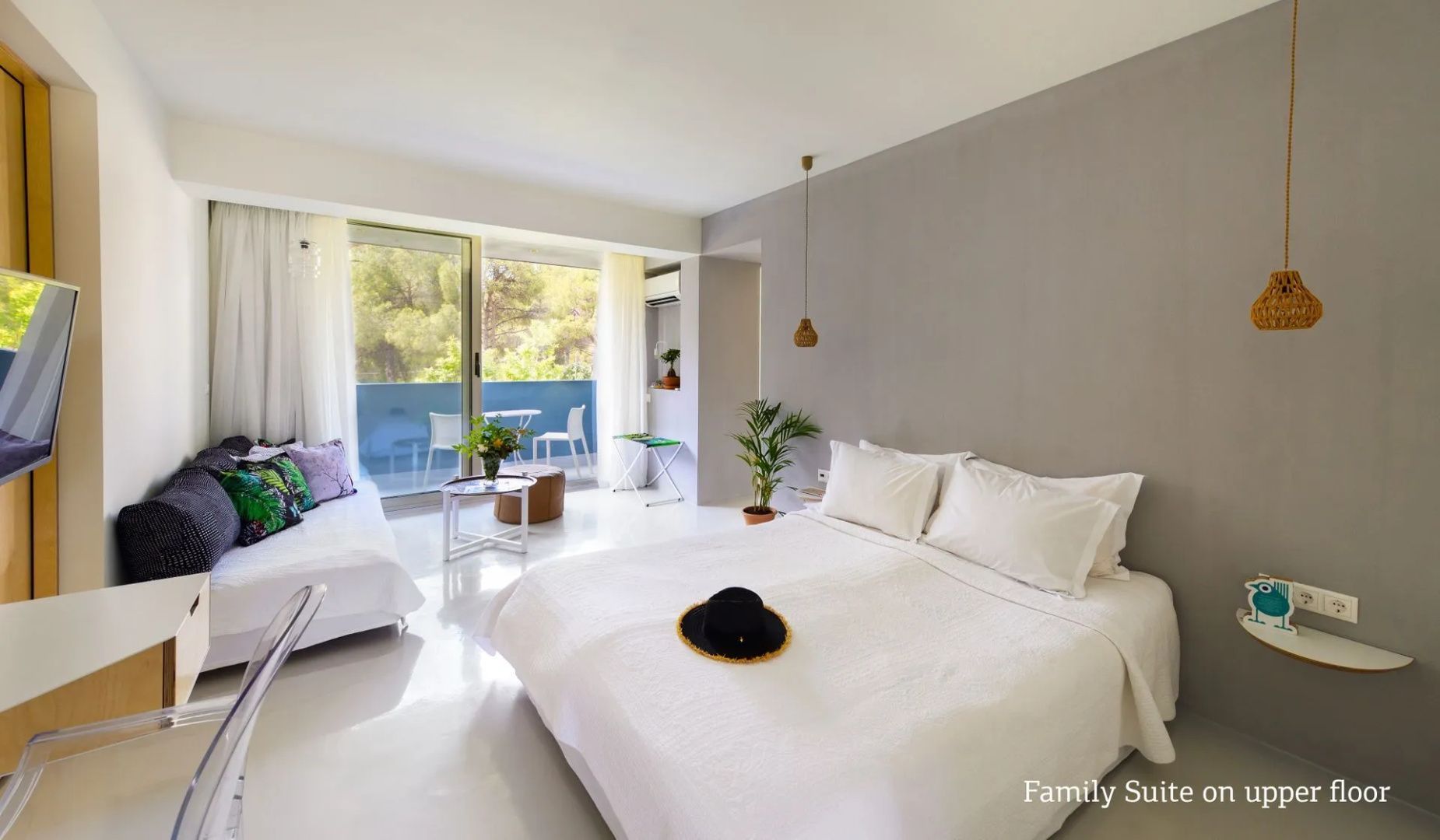 Doppelbett mit Hut im Vordergrund und helles Zimmer mit Sitzgelegenheit vor dem Balkon.