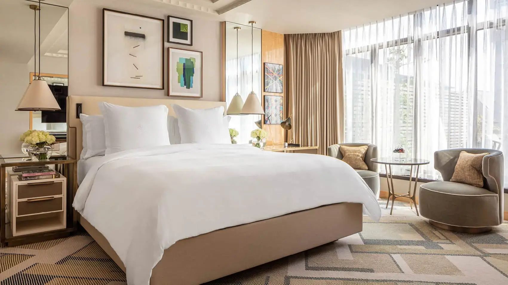 Hell eingerichtetes Hotelzimmer mit einem großen Doppelbett, Wandbildern und einem Sessel am Fenster.