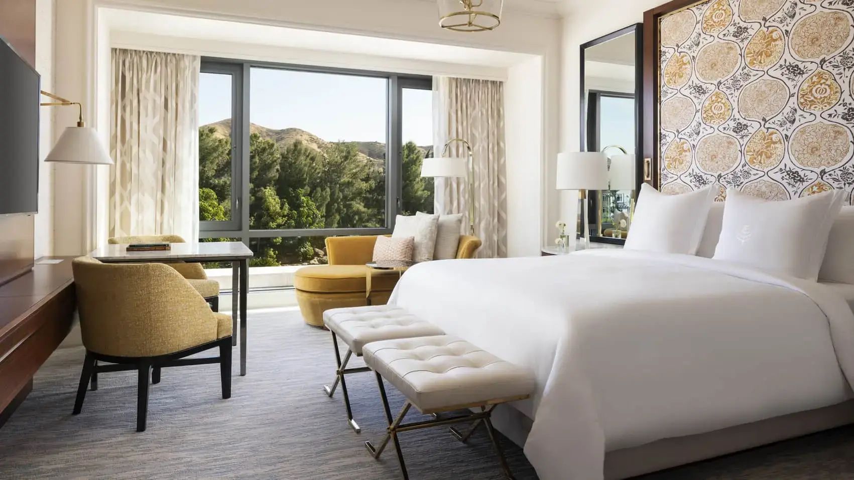 Helles Hotelzimmer mit großem Doppelbett, gelber Récamiere und Blick auf grüne Hügel.