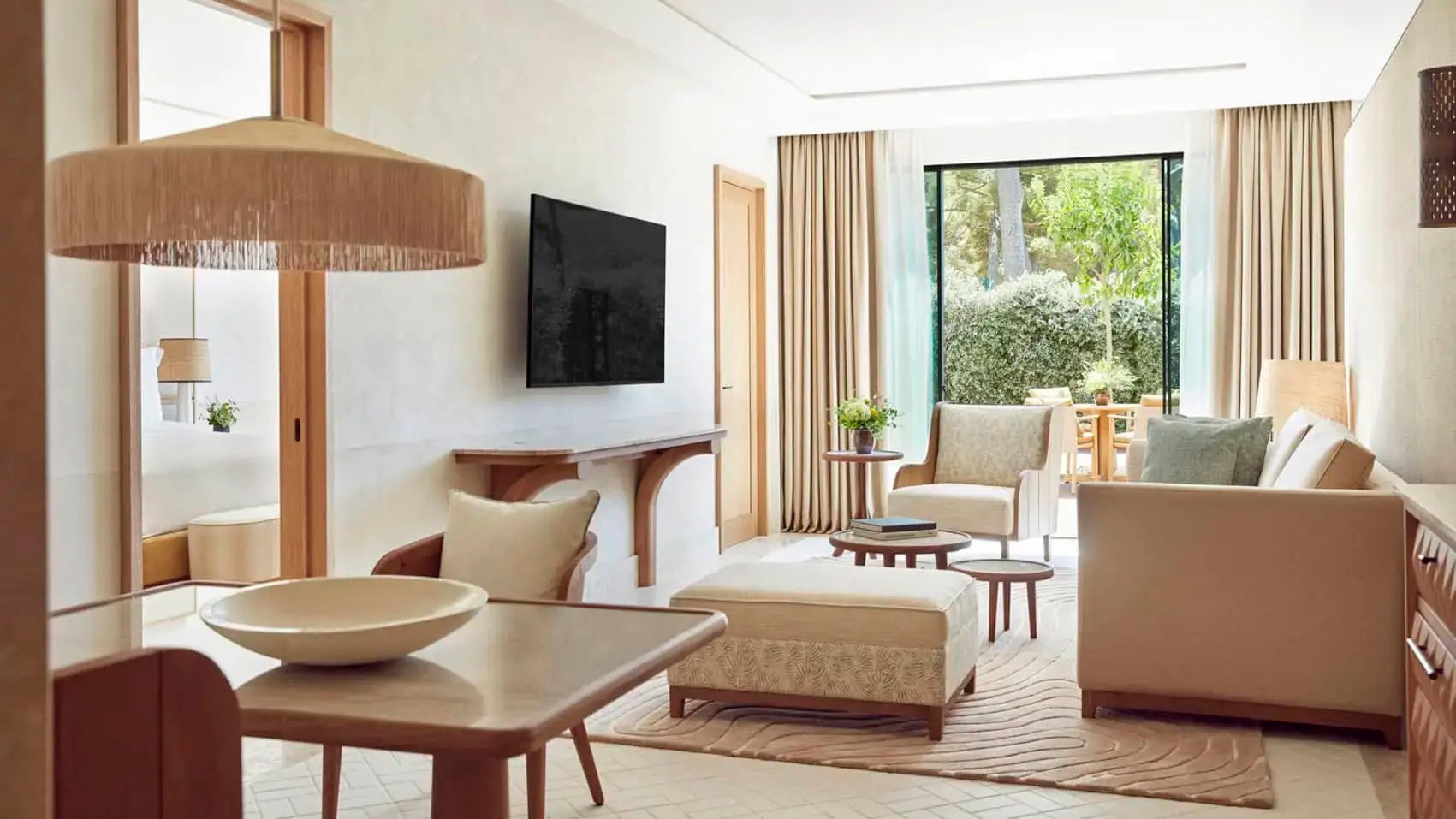 Heller Wohnbereich einer Suite mit Sessel, Hocker, TV und Blick in den grünen Garten.