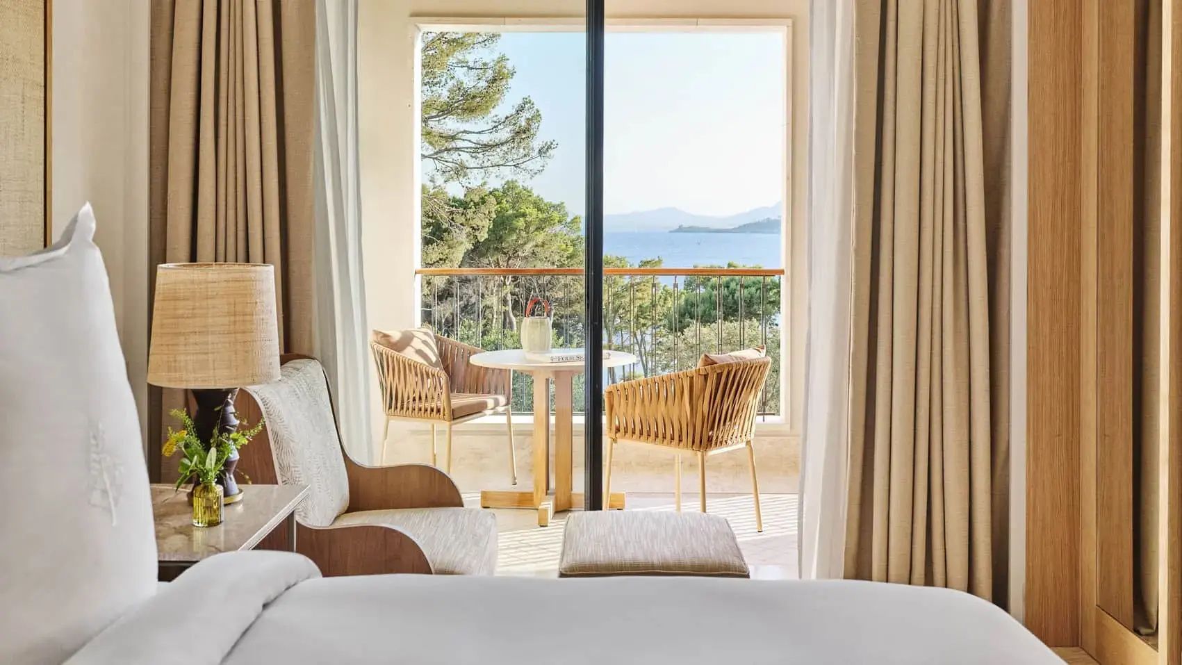 Blick vom Zimmer über die Balkonmöbel auf Bäume und das blaue Meer.