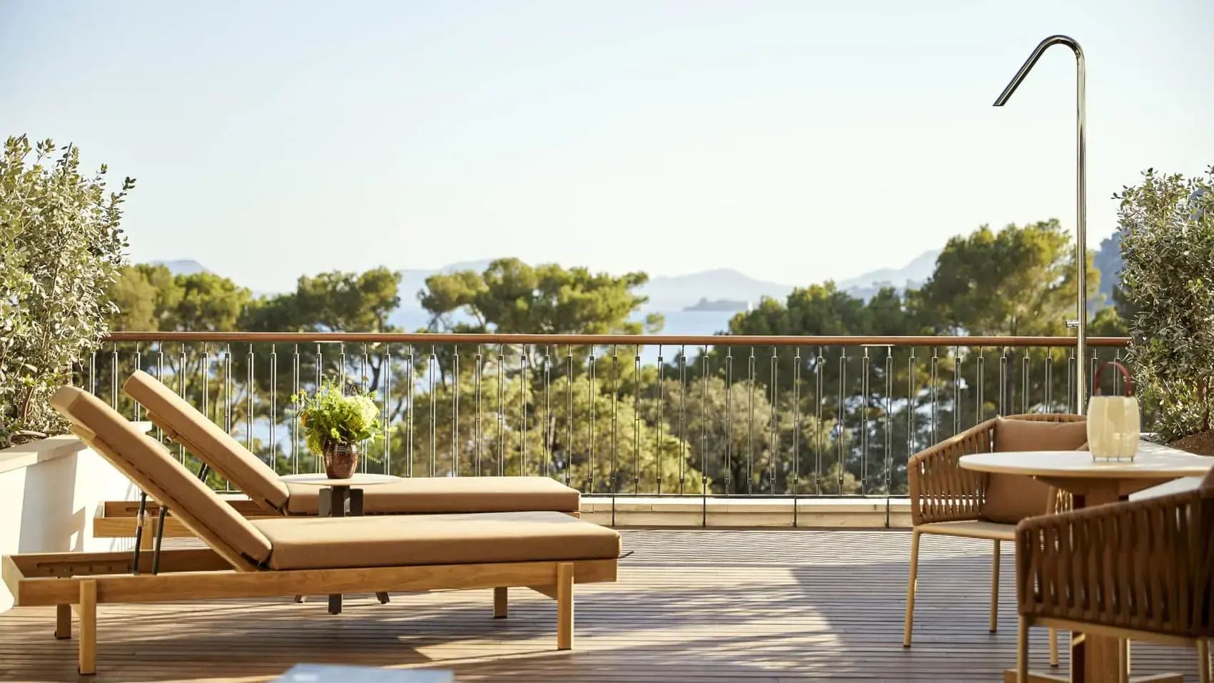 Holzliegen auf einer Terrasse mit Geländer und Ausblick auf grüne Pinien und das Meer.