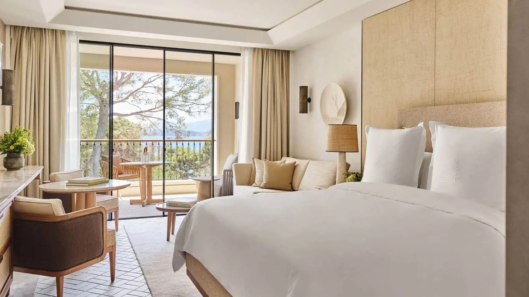 Heller Hotelraum mit weiß bezogenem Bett, Sofa und Zugang zum Balkon mit Blick auf Bäume und das Meer.