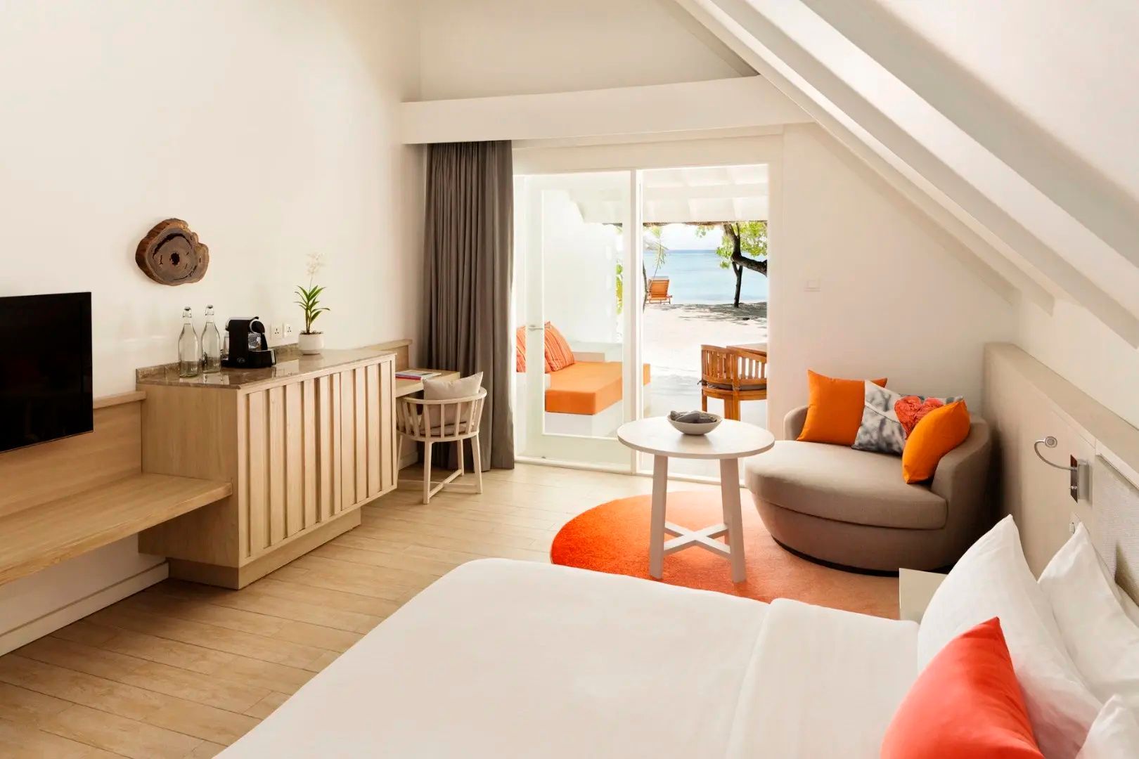 Hell eingerichtetes Zimmer mit orangefarbenen Kissen, Sessel und Blick auf den Strand.