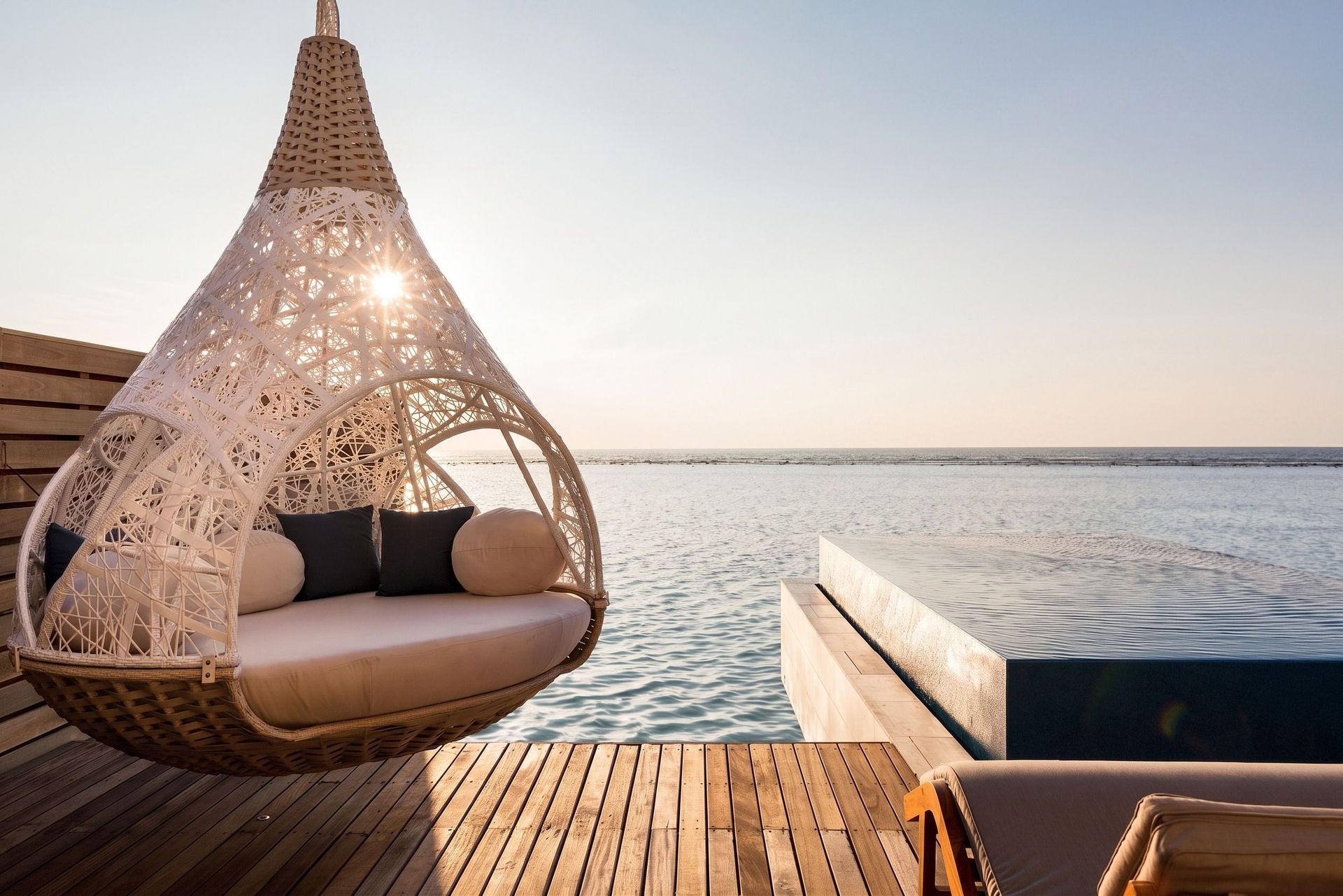 Geflochtener Hängesessel mit Kissen neben einem Pool. Blick auf das Meer.