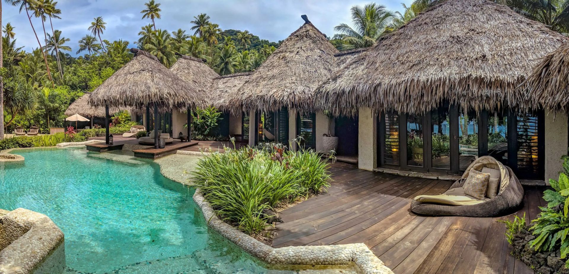 Resort-Villen mit traditionellen Strohdächern an einer Holzterrasse mit Pool und Bepflanzung.