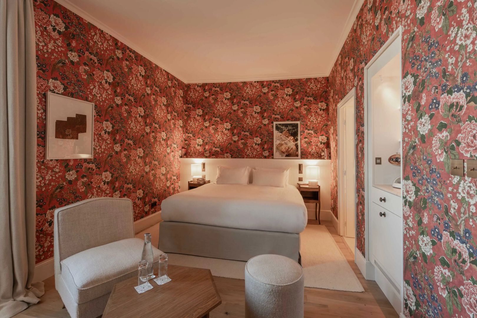 Hotelzimmer mit roter Blumentapete, Doppelbett, Holztisch und bequemem Sessel.