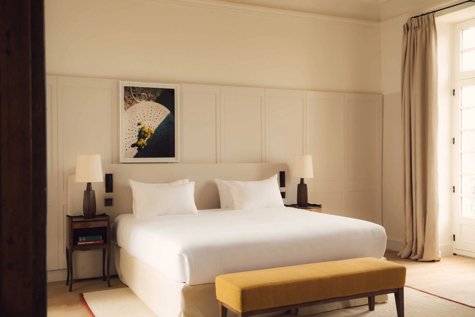 Doppelbett mit weißer Bettwäsche, gelber Sitzbank und Wandpaneelen im Hotelzimmer.