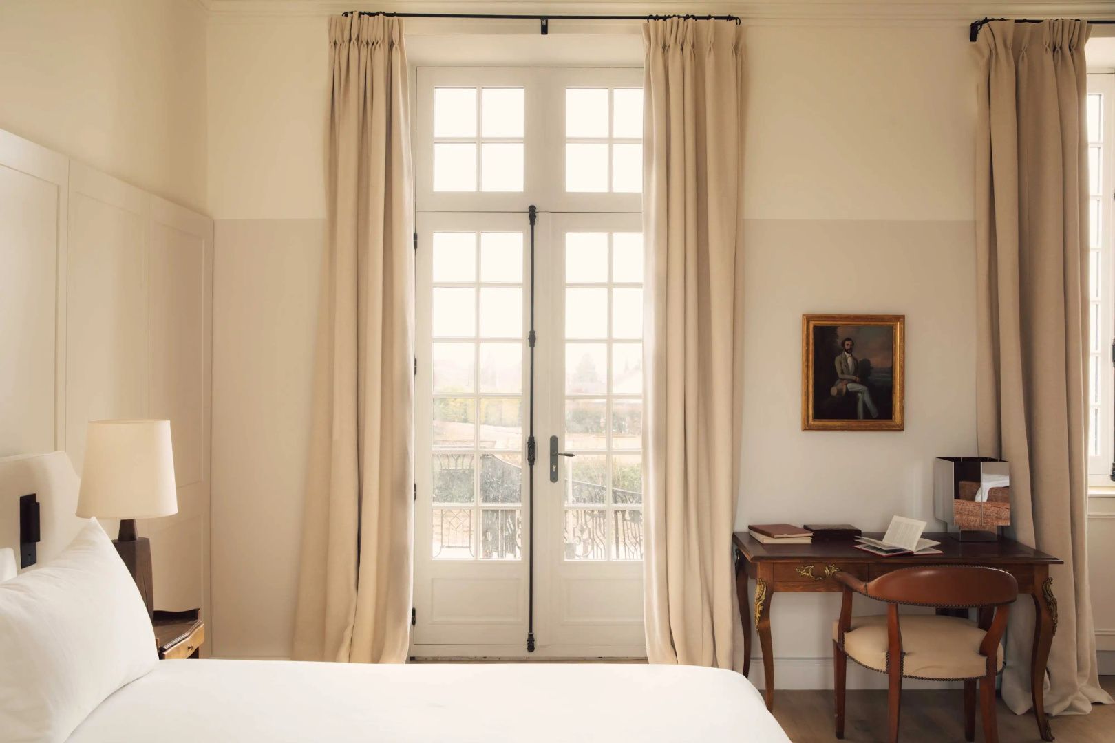 Bodentiefes Fenster mit beigen Vorhängen und einem Holzschreibtisch im Hotelzimmer.