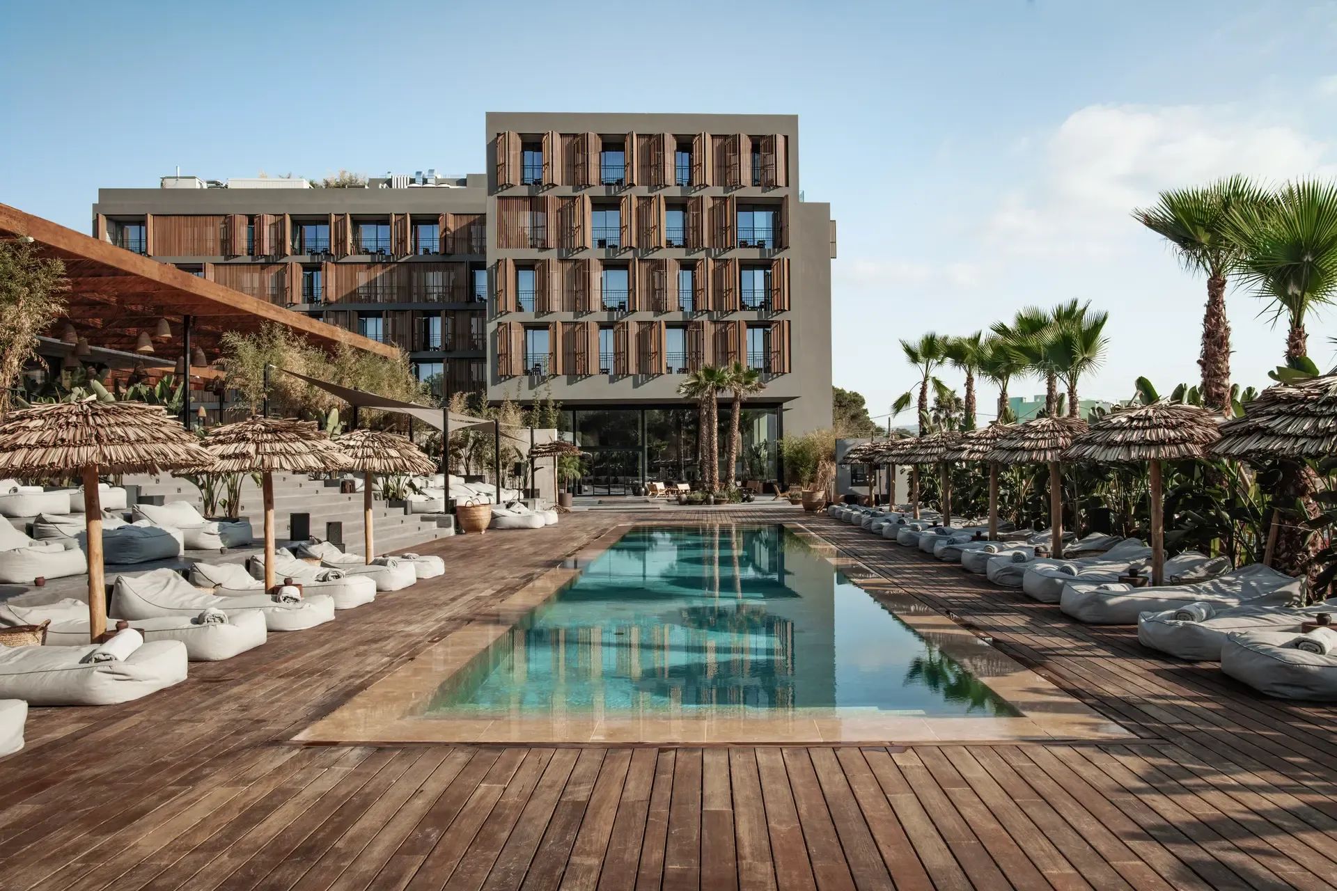 Poolbereich des OKU Ibiza mit minimalistischer Architektur, Naturmaterialien, Sonnenliegen mit Strohdächern und entspanntem Boho-Design vor dem modernen Hotelgebäude.