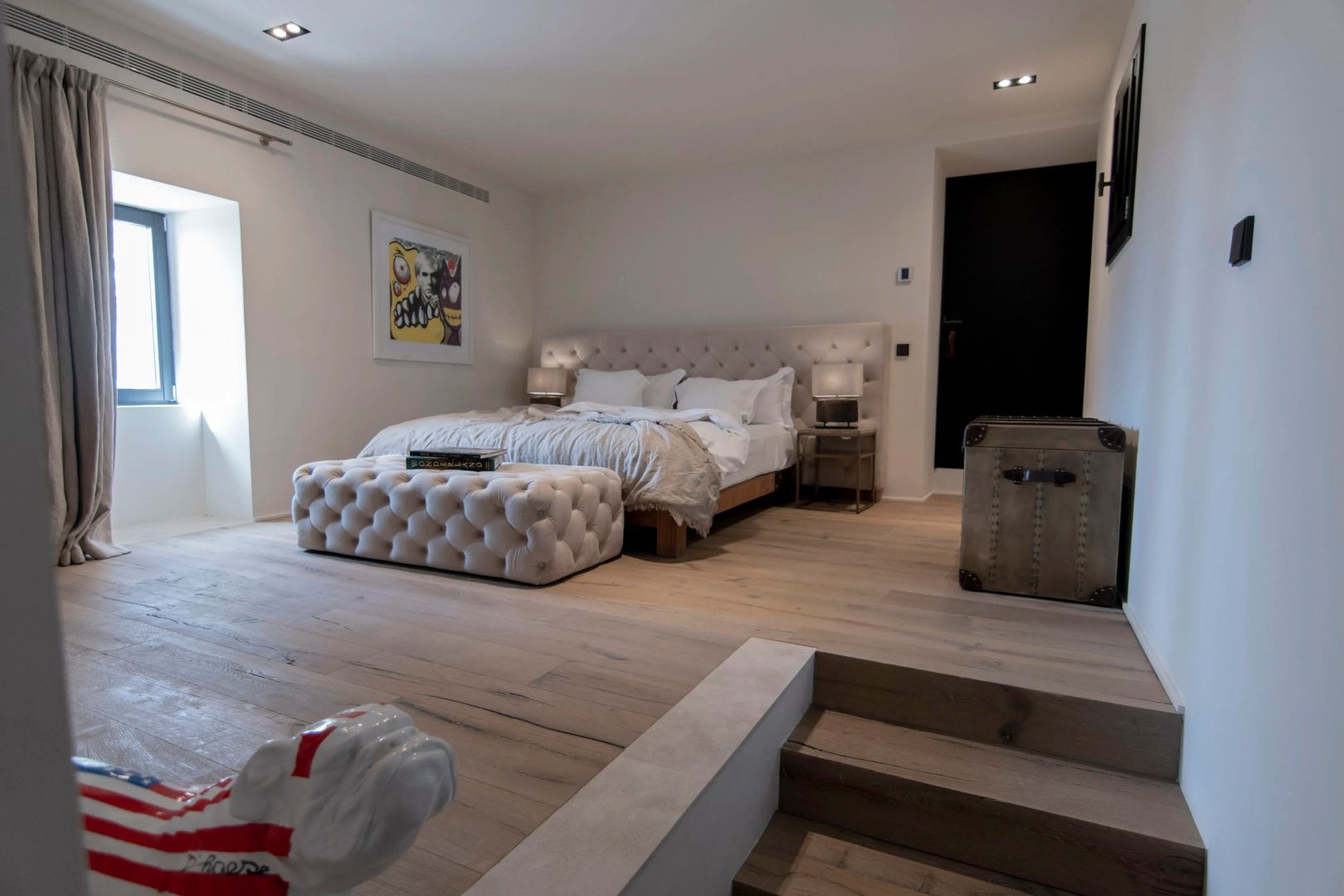 Hell eingerichtetes Schlafzimmer mit großem Polsterbett, Holzboden und gepolsterter Sitzbank.