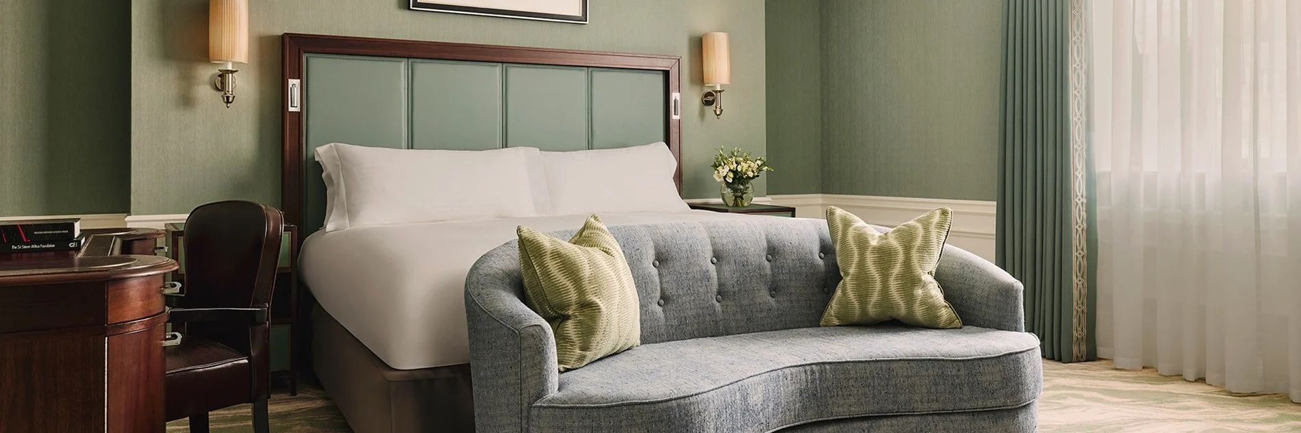 Hellgrünes Hotelzimmer mit grauem Sofa im Vordergrund und weiß bezogenem Doppelbett dahinter.