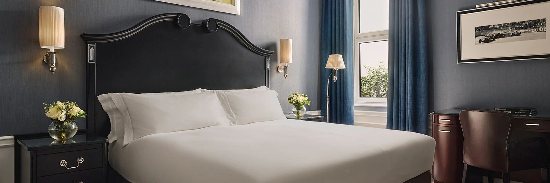 Doppelbett mit weißer Bettwäsche, dunklem Kopfteil und einer Wandlampe im Hotelzimmer.