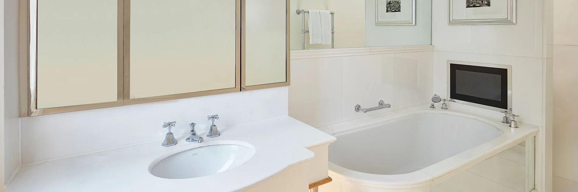 Hell gefliestes Badezimmer mit weißem Waschbecken, Spiegelschrank und der Kante einer Badewanne.