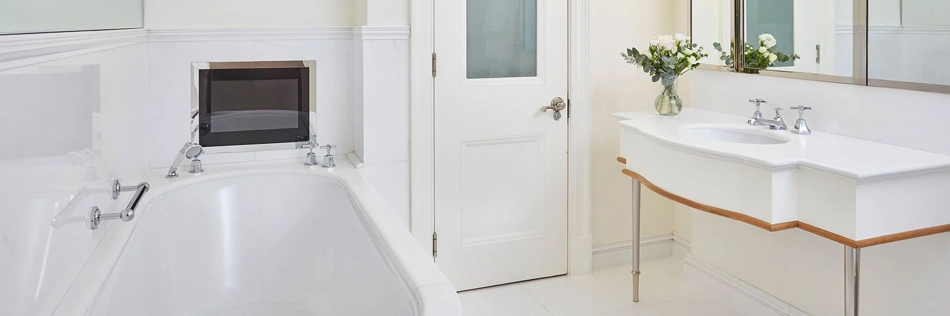 Weiße Badezimmertür mit Glaseinsatz neben flach eingebauter Badewanne und hellen Fliesen.
