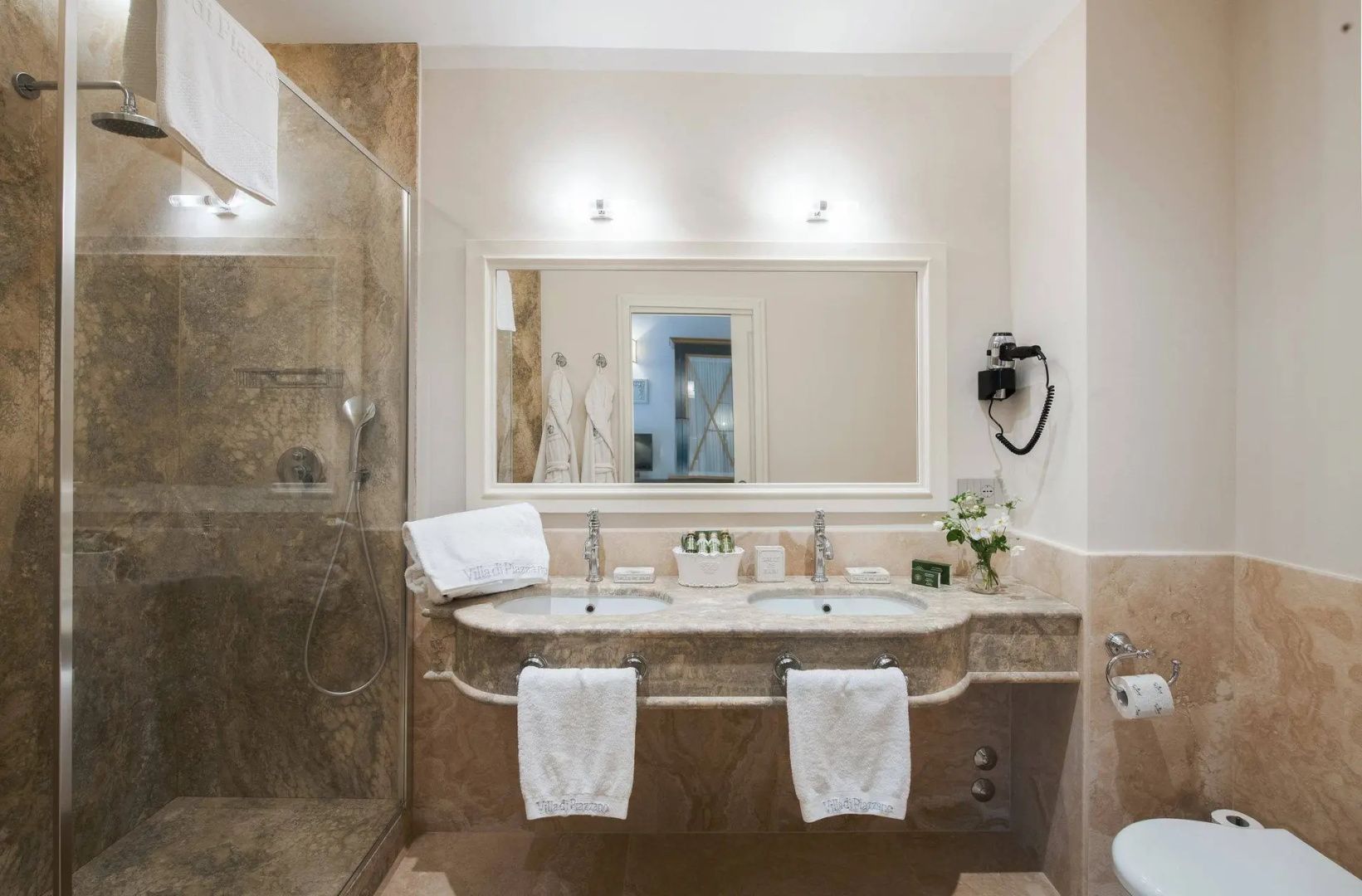 Badezimmer mit Doppelwaschtisch aus Naturstein, großem Spiegel und einer Dusche aus Glas.