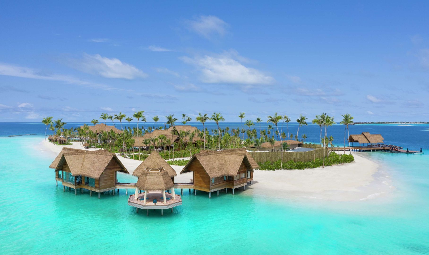 Wasser-Villas mit Strohdächern an einer türkisfarbenen Lagune mit Sandstrand und Palmen.