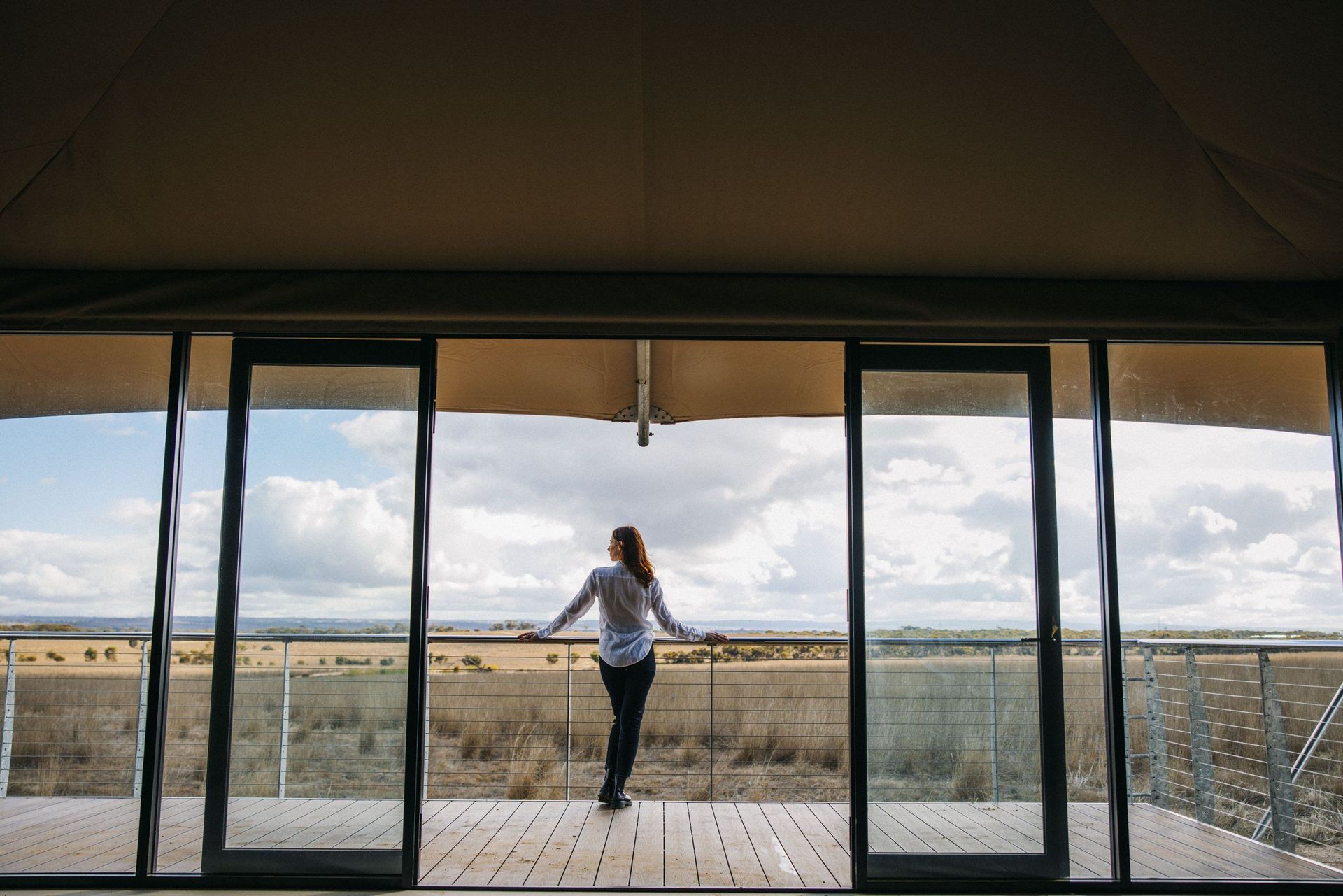 Glasfront eines Safari-Lodge-Zelts, auf dem Deck blickt eine Frau über das Grasland