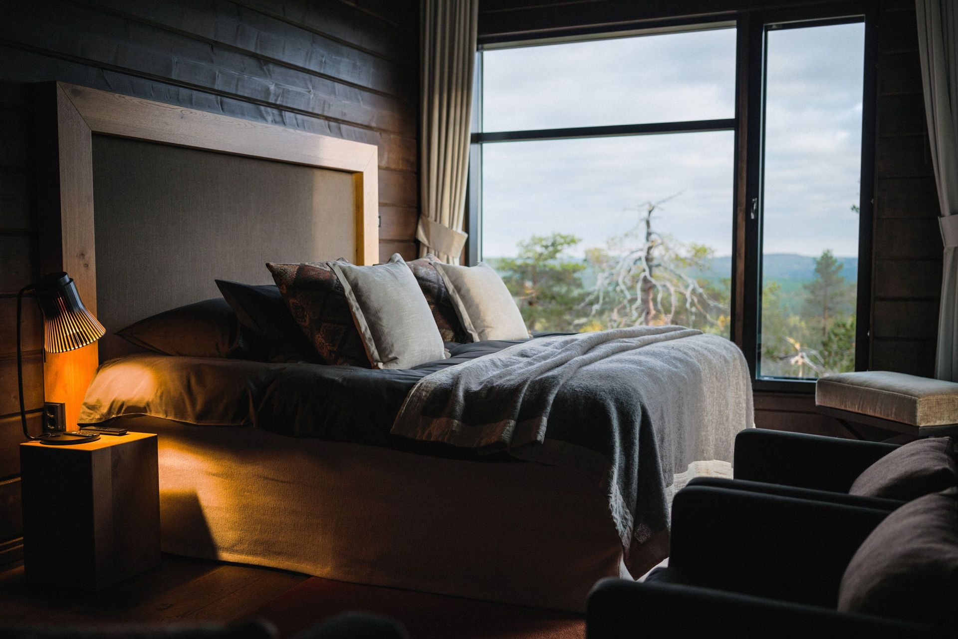 Schlafzimmer der Octola Lodge mit Doppelbett, Leselampe und Fenster zum Kiefernwald im Herbst