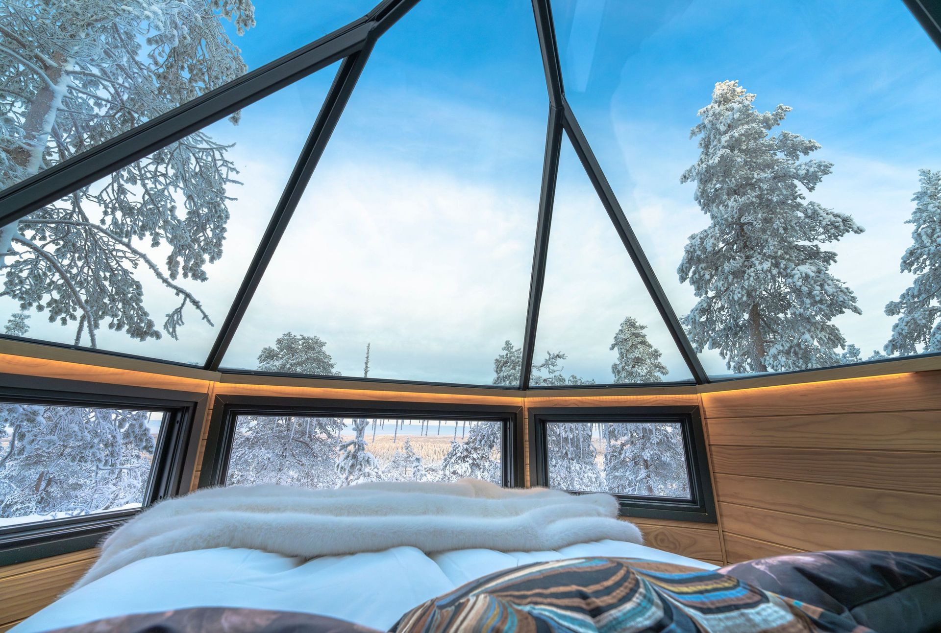 Bett unter dem Glasdach der Glass Igloo Suite, darüber verschneite Kiefern und blauer Himmel