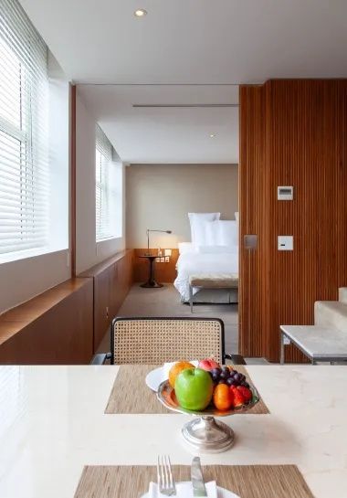 Blick über einen Esstisch mit Obstschale hin zu einem hellen Doppelbett.