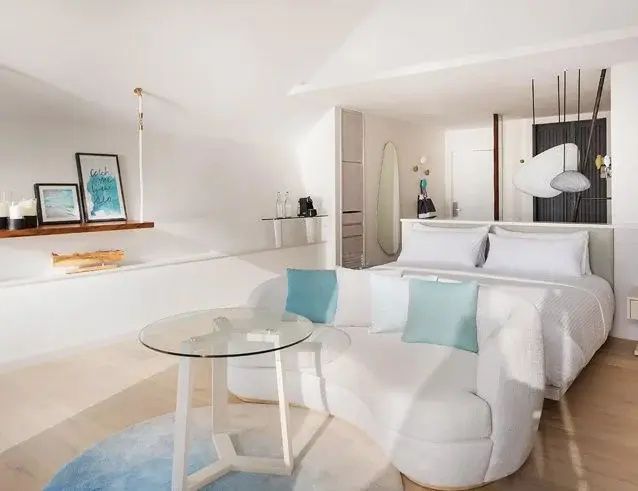 Helles Hotelzimmer mit weißem Sofa, Glasstisch und Doppelbett im Hintergrund.
